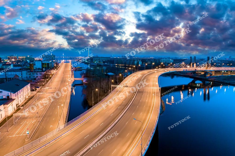 На фотографии изображён вантовый мост Санкт-Петербурга в вечернее время. Магистраль, освещённая яркими фонарями, плавно изгибается над рекой, отражая свет в её спокойных водах. Вдали видны огни города и современные здания, создавая динамичную городскую картину на фоне закатного неба.